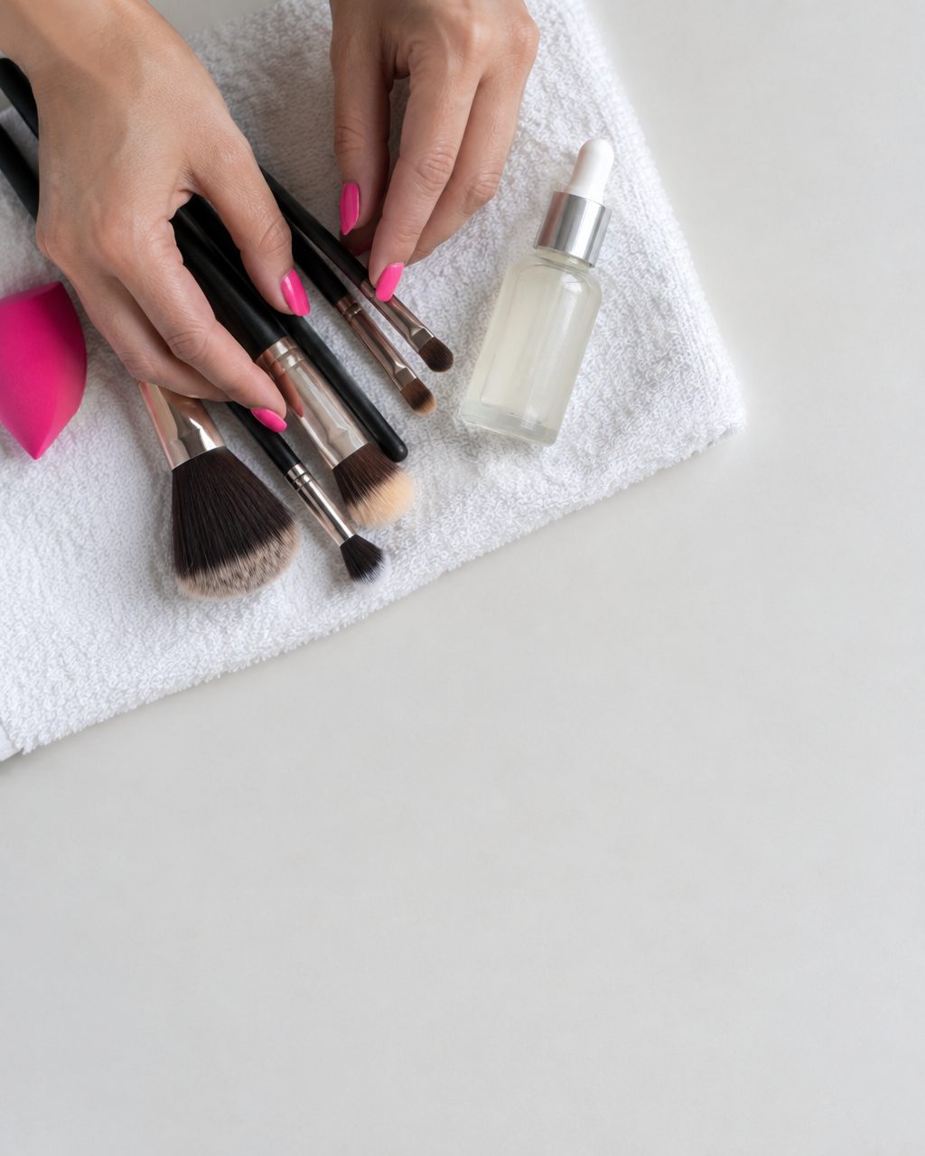 Mãos com esmalte rosa organizando pincéis de maquiagem e um sérum.